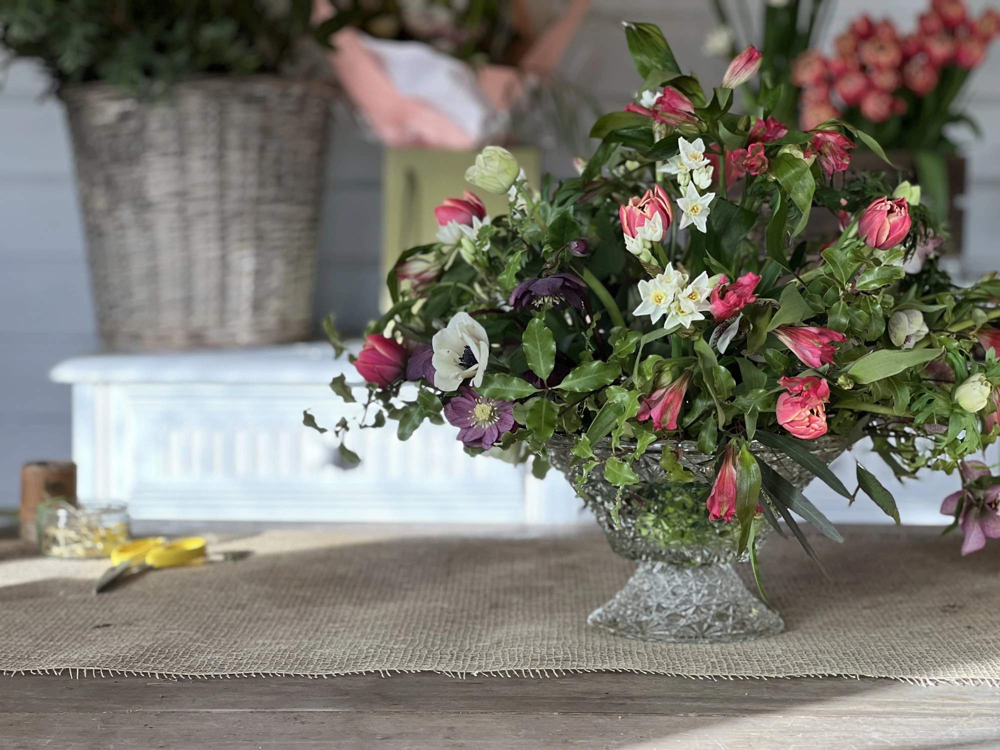 A crystal footed compote arrangement of pink tulips, purple hellebores, narcissus and anemones on a burlap runner — hand-designed by Carol, entirely foam-free.